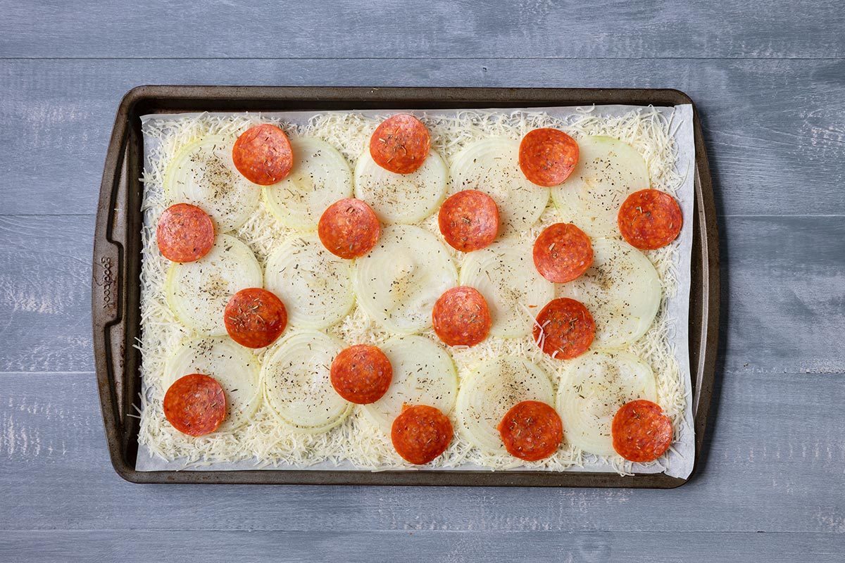 Unbaked onion "pizza" with cheese and pepperoni layered on top of sliced onions on a baking sheet, on top of a gray table