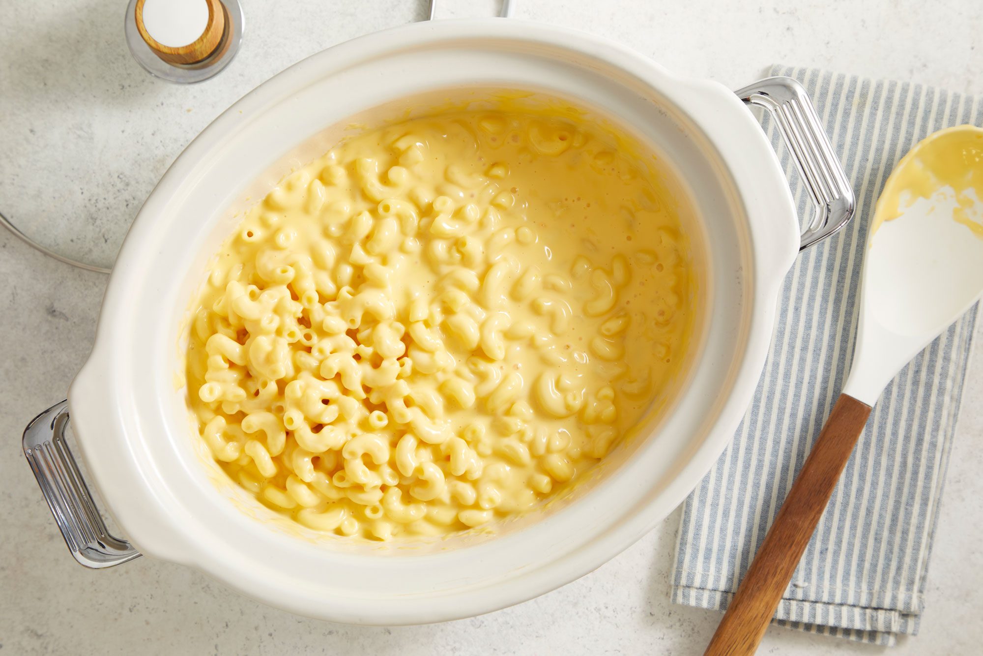 overhead shot of a dish of Potluck Macaroni and Cheese presented in a glossy white serving dish; beside the dish, there is a spoon with a wooden handle resting on a light blue and white striped napkin, the background is a light, neutral surface