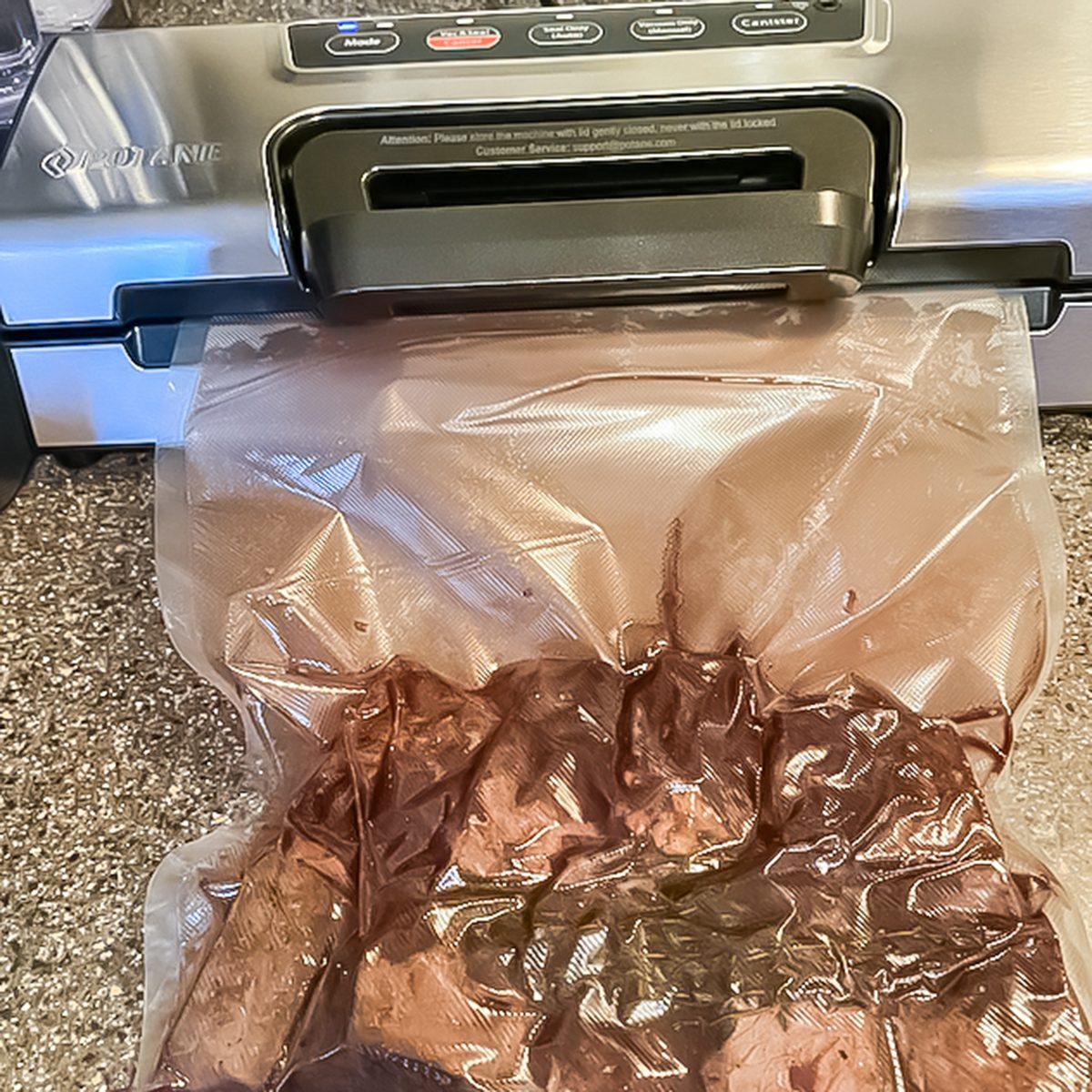 A vacuum sealing machine on a kitchen counter seals a plastic bag containing pieces of dark-colored meat or food. The bag is partially inside the machine, which has buttons and labels on its front panel.