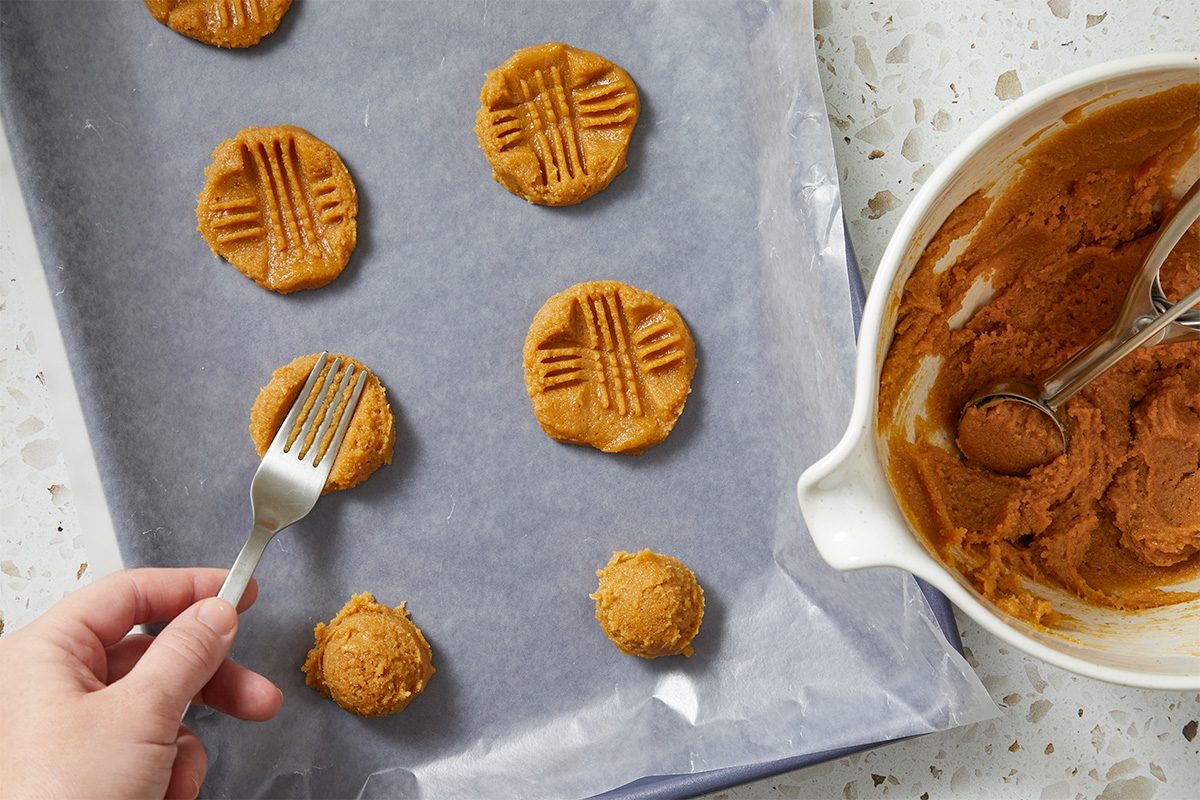 A hand presses a fork into peanut butter cookie dough balls on a parchment-lined baking sheet, creating a crisscross pattern. A mixing bowl with dough and a scoop sit nearby.