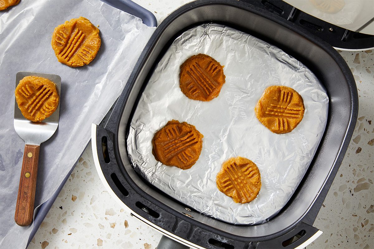 Air fryer basket lined with foil holding four unbaked peanut butter cookies with fork marks on top. Next to it, a baking sheet with three more cookies and a spatula with a wooden handle.