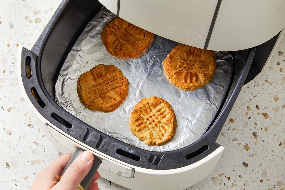 A hand pulls out an air fryer drawer lined with foil, containing four baked peanut butter cookies with a crisscross pattern on top.