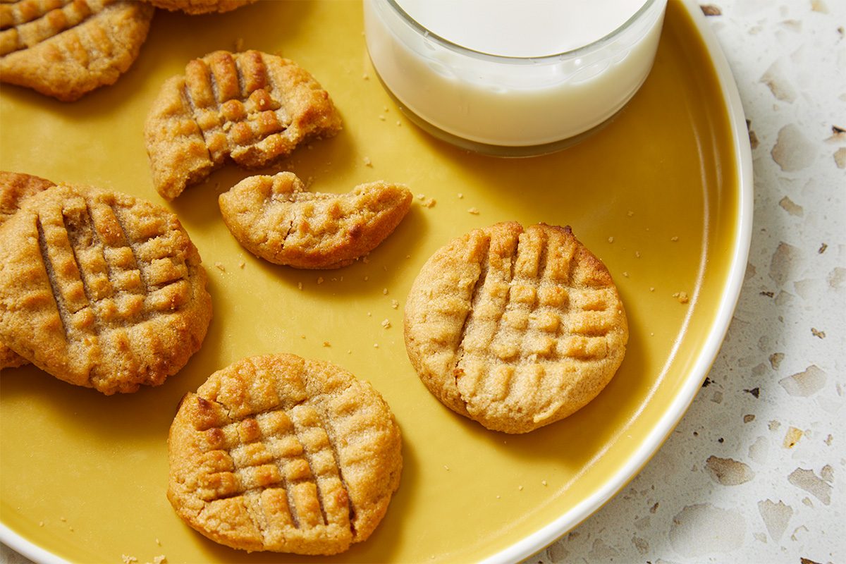 A yellow plate with several peanut butter cookies, some whole and one partially eaten, next to a glass of milk. The cookies have a crisscross pattern on top. The plate is on a light, textured surface.