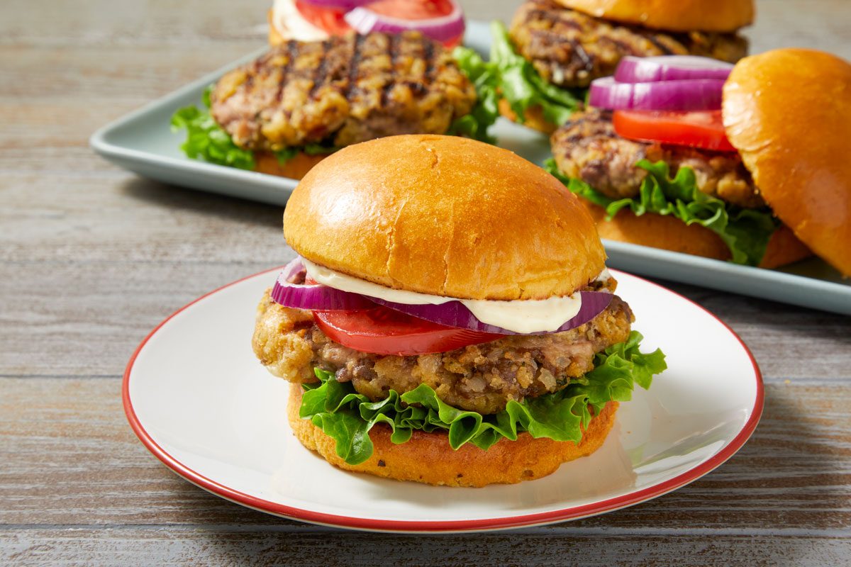 3/4th shot of Beef and Pork Burger displayed in the foreground on a white plate with a red rim; in the background, additional burgers can be seen resting on a rectangular plate, the setting features a rustic wooden table