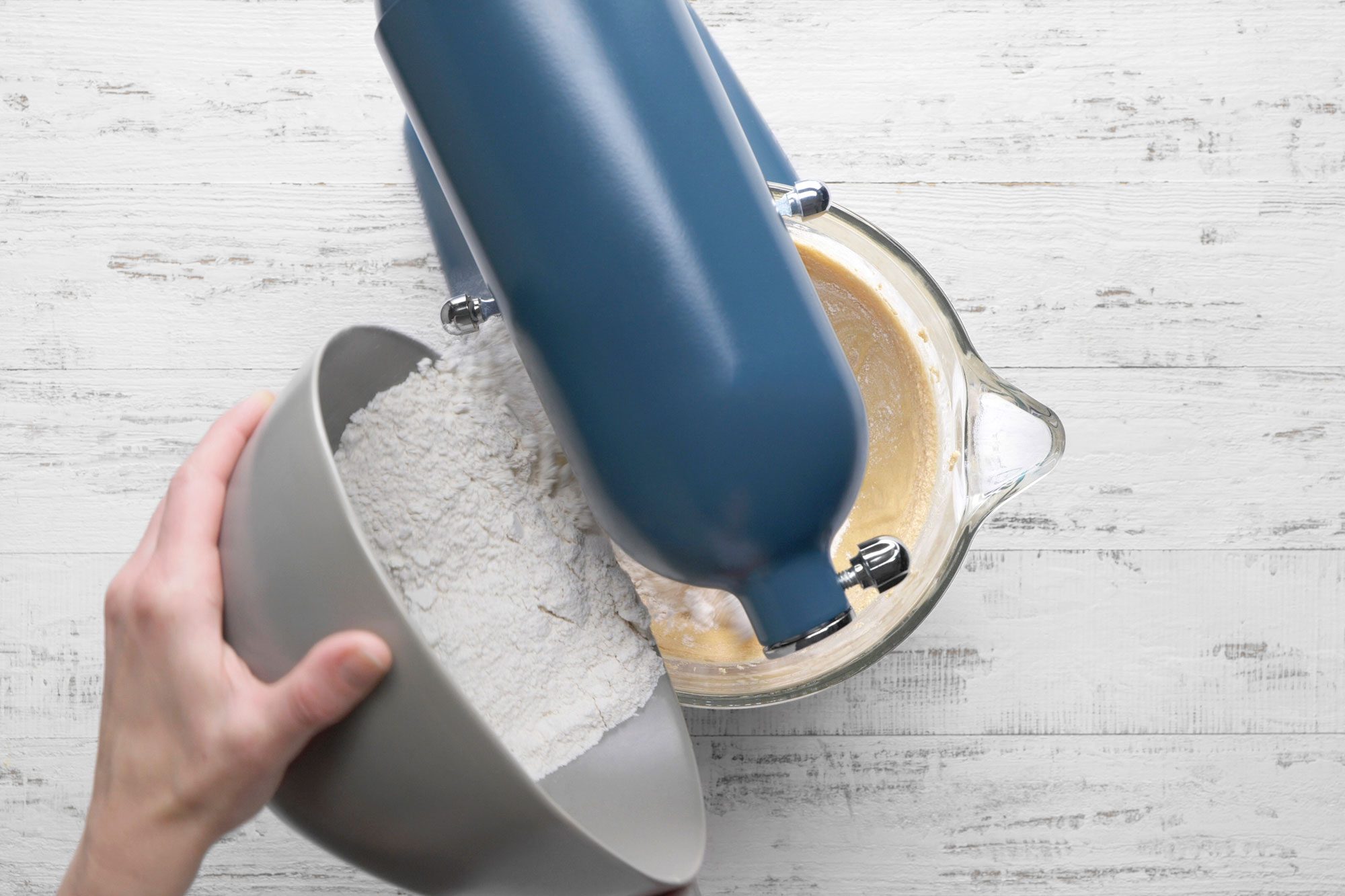 overhead shot of a hand pours flour from a gray bowl into a stand mixer with blue housing, which contains a yellow batter, on a white wooden surface