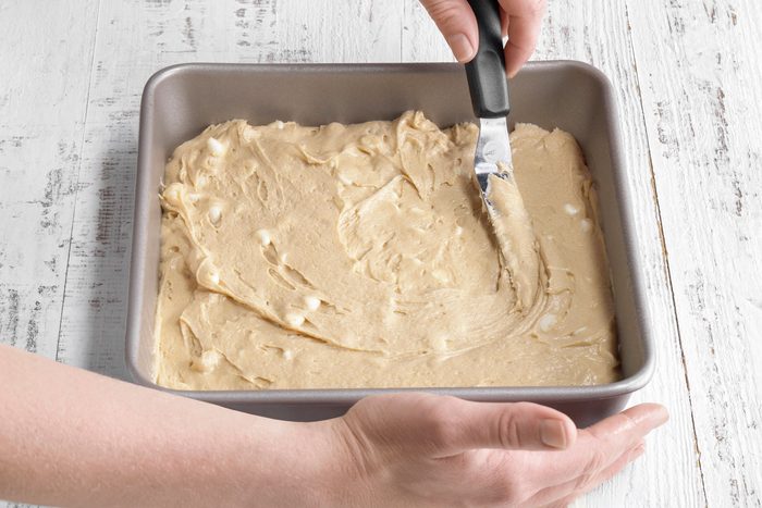 3/4th shot of a person spreads thick batter evenly in a square metal baking pan using a spatula on a white wooden surface