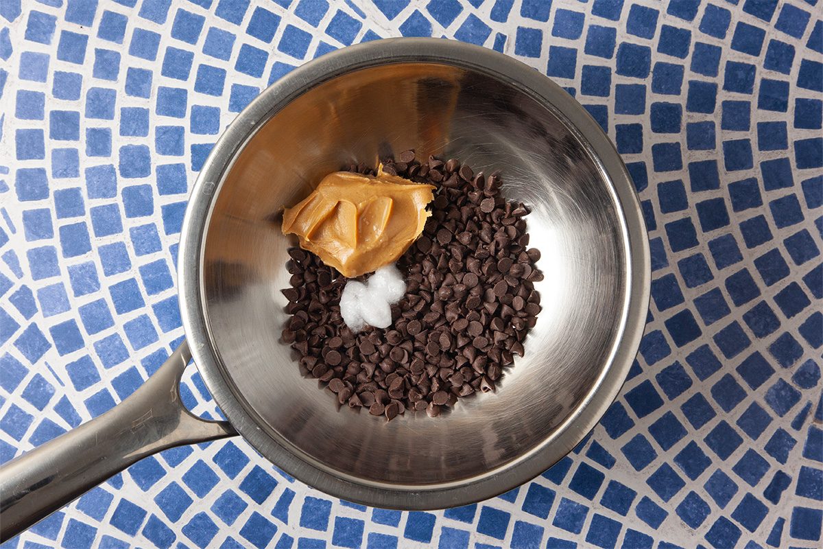 A metal saucepan on a blue and white mosaic surface contains chocolate chips, a dollop of peanut butter, and a small amount of coconut oil.