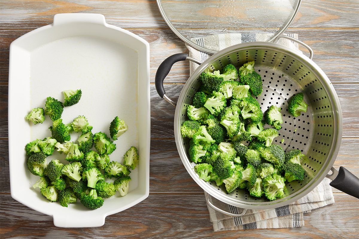 Broccoli florets are scattered in a white baking dish on the left, while more broccoli is in a metal colander inside a pot with a glass lid on the right, all placed on a wooden surface.
