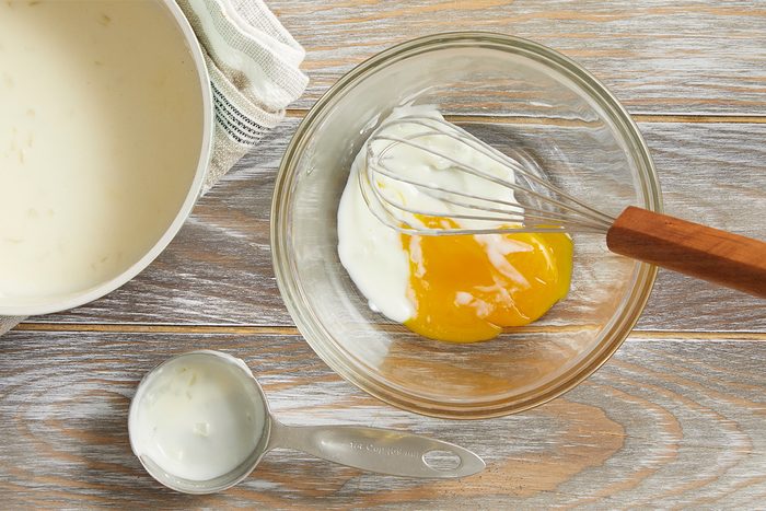 A glass bowl with two egg yolks and white yogurt being whisked together, next to a measuring cup with yogurt, a dish towel, and a white bowl on a wooden surface.