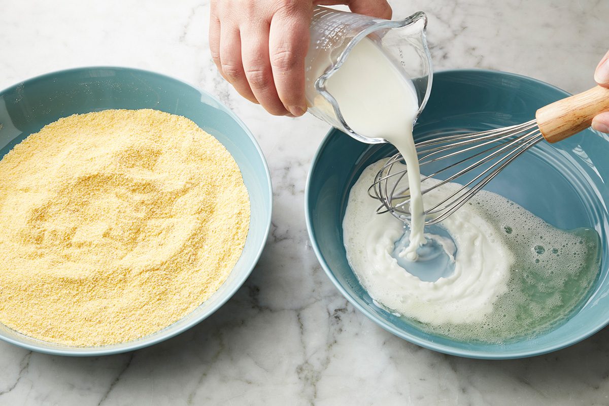A person pours milk from a small pitcher into a blue bowl while whisking, next to another blue bowl filled with cornmeal on a marble countertop.
