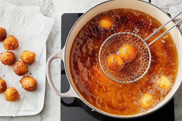 A pot of oil on a stovetop with golden-brown balls being fried; a slotted spoon holds some above the oil, while more finished balls rest on paper towels nearby.