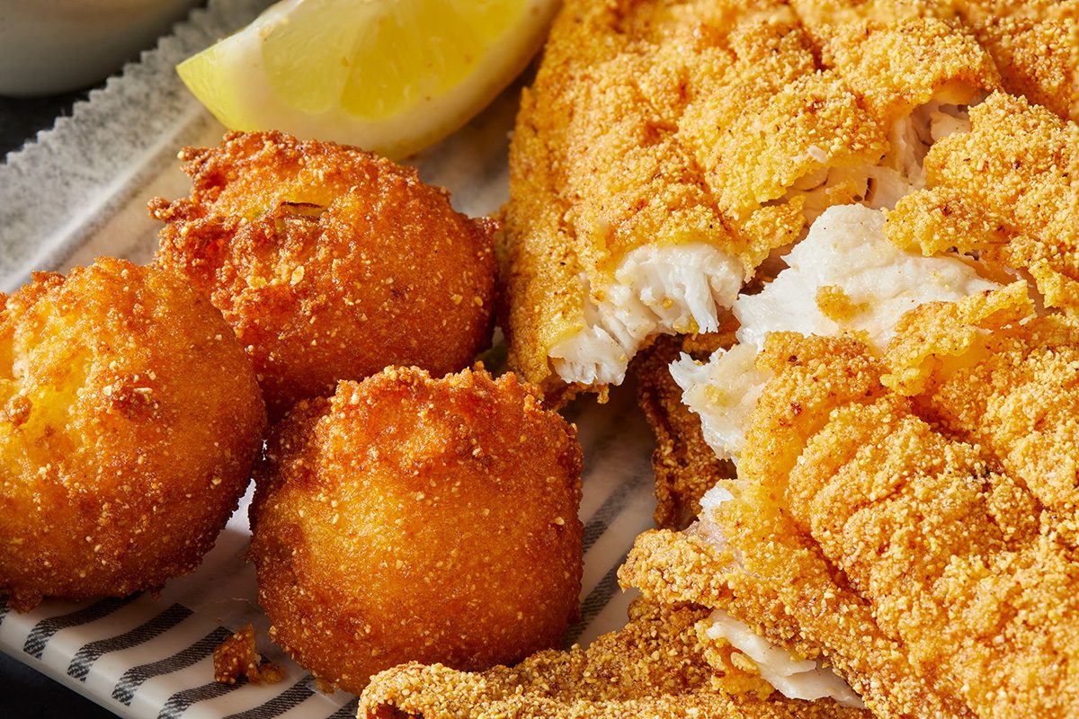 Close-up of golden fried catfish fillets with a crispy cornmeal coating, served alongside two hush puppies and a wedge of lemon on a plate.