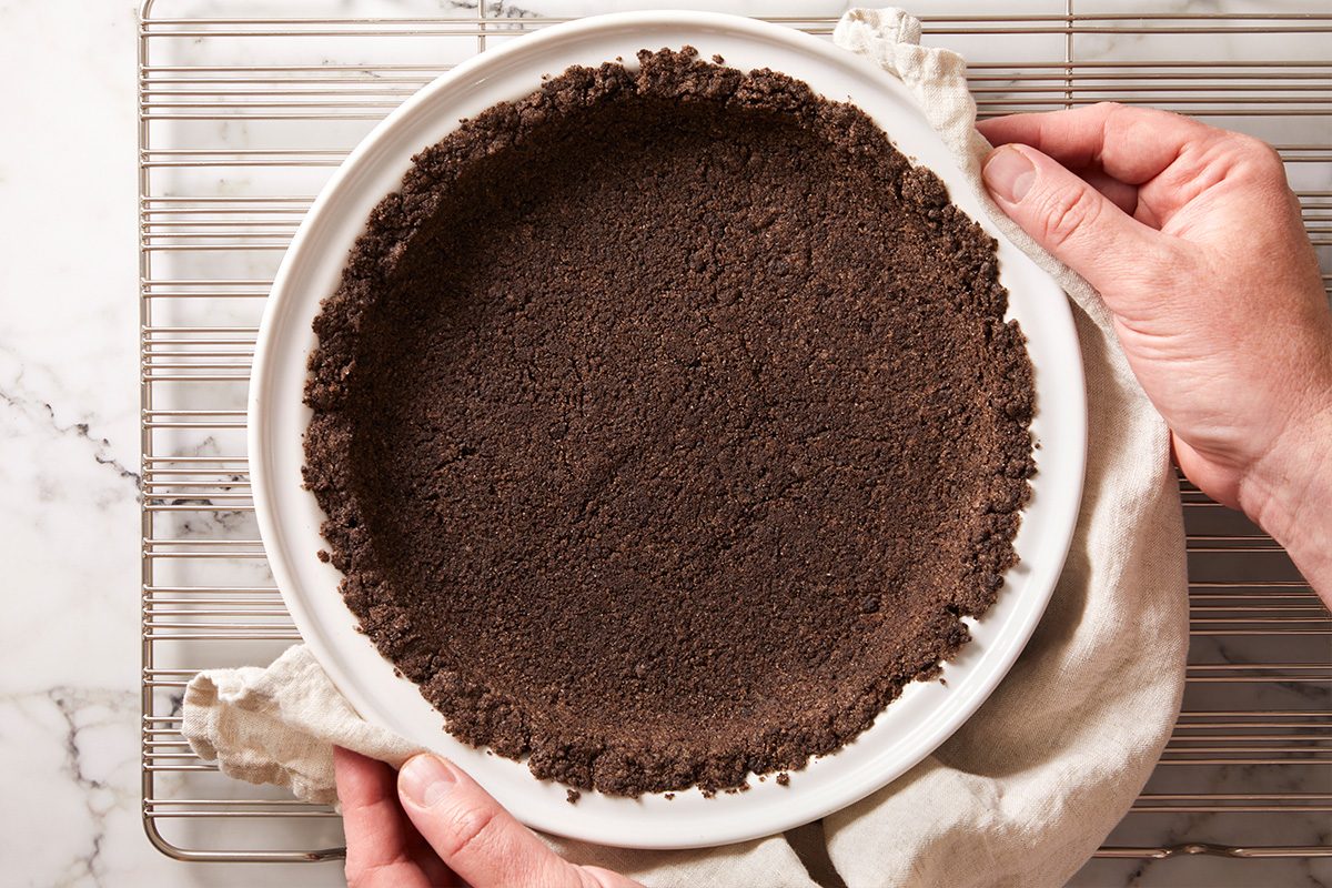 A person's hands holding a white pie dish with a chocolate cookie crumb crust, resting on a cooling rack atop a marble surface.
