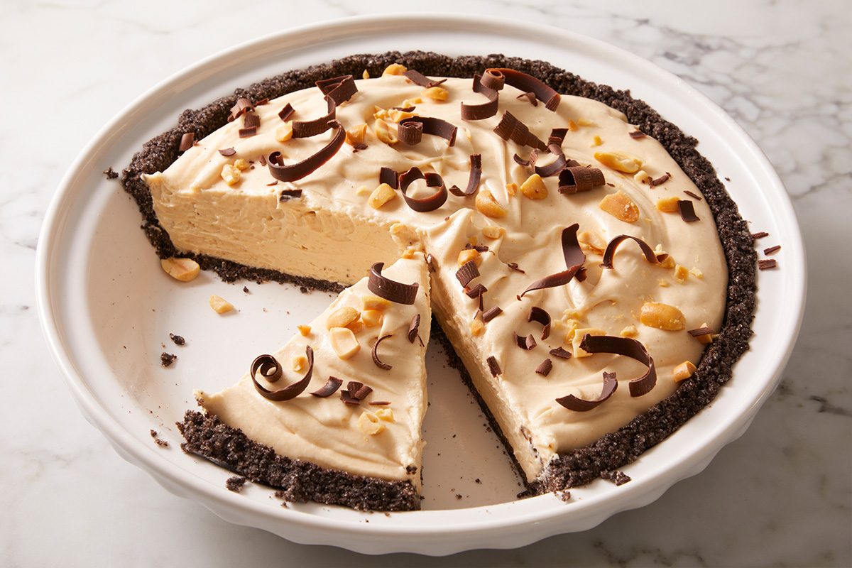A creamy peanut butter pie with a chocolate cookie crust in a white pie dish. One slice has been removed, and the pie is topped with chocolate curls and chopped peanuts.
