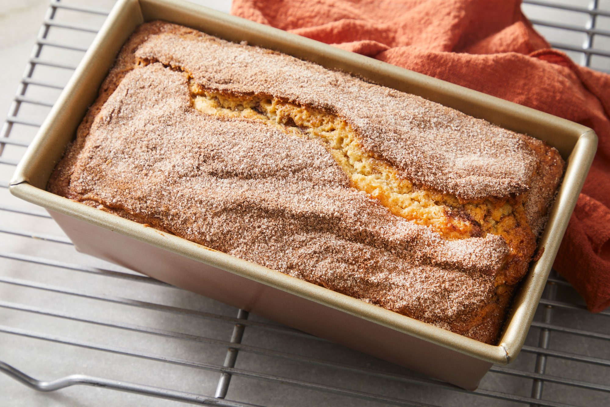 overhead shot of a freshly baked cinnamon sugar bread loaf sits in a rectangular metal pan, resting on a cooling rack with an orange cloth nearby, The loaf has a cracked top and a generous dusting of cinnamon sugar