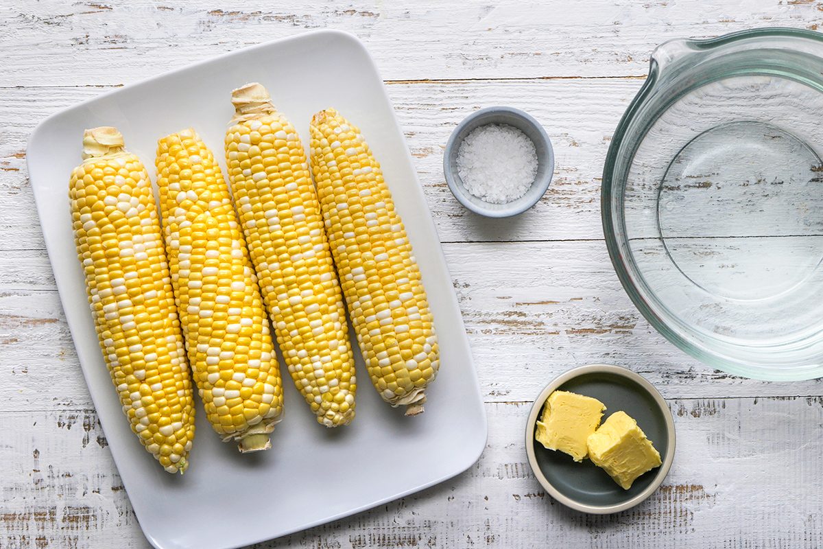 Four ears of raw corn on a white plate next to a small bowl of salt, a dish with two pats of butter, and a glass bowl of water on a white wooden surface.
