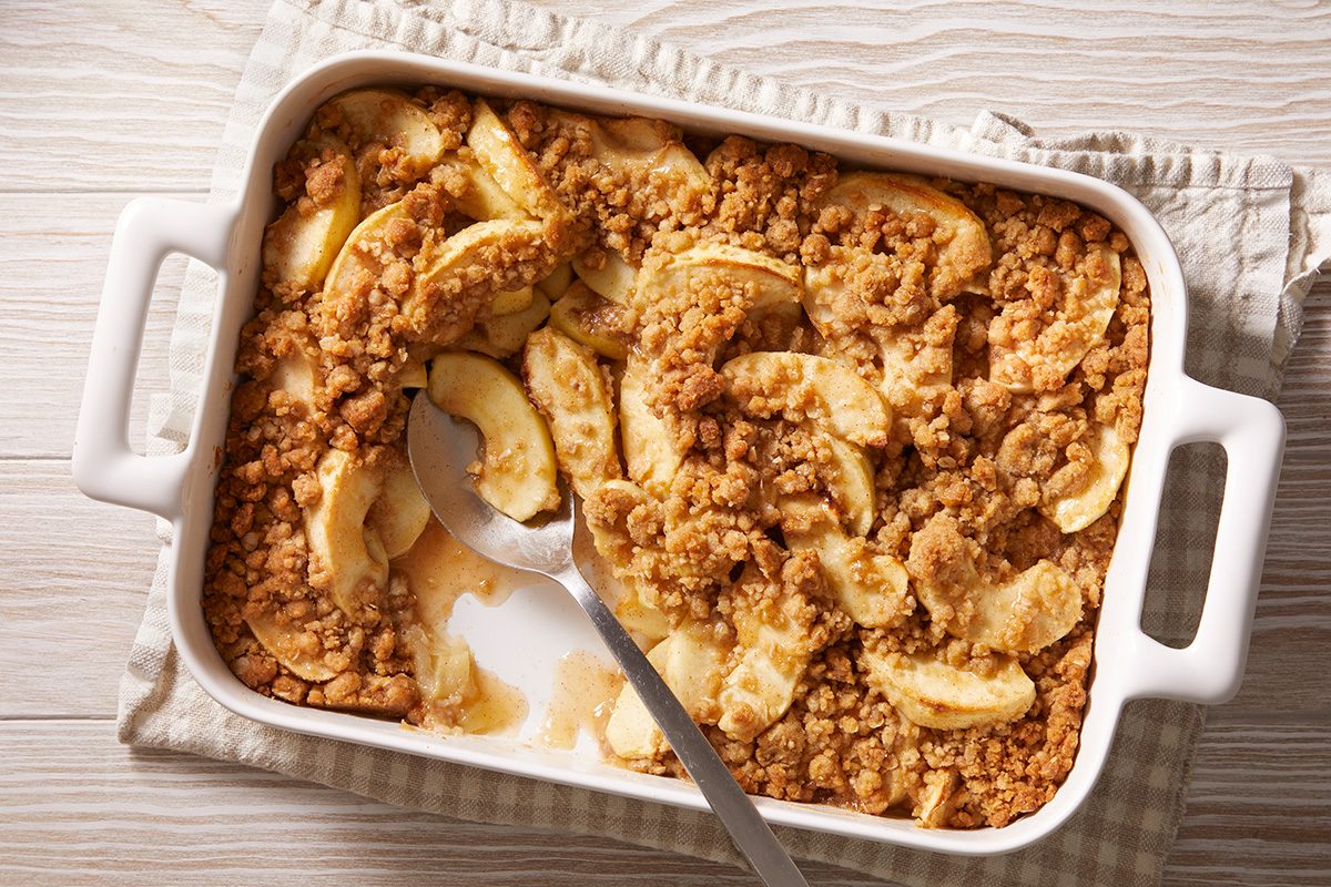 A rectangular white baking dish filled with apple crisp, featuring baked apple slices topped with a golden brown crumbly oat topping. A serving spoon rests inside, with some crisp scooped out.
