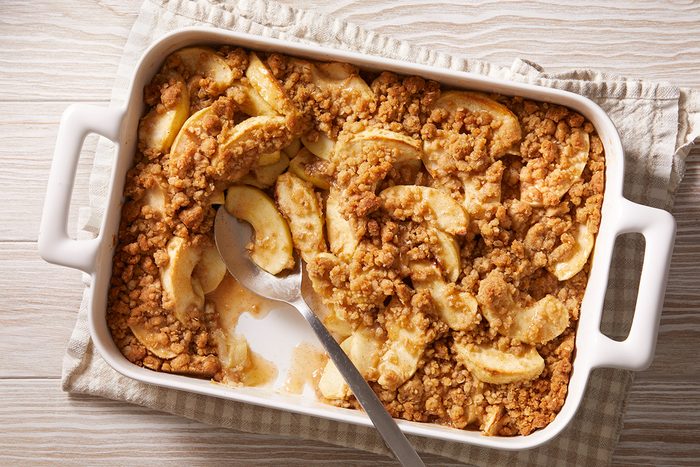 A rectangular white baking dish filled with apple crisp, featuring baked apple slices topped with a golden brown crumbly oat topping. A serving spoon rests inside, with some crisp scooped out.