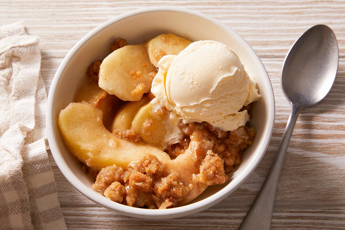 A white bowl filled with apple crisp topped with a scoop of vanilla ice cream, placed on a light wooden surface next to a spoon and a beige striped cloth.