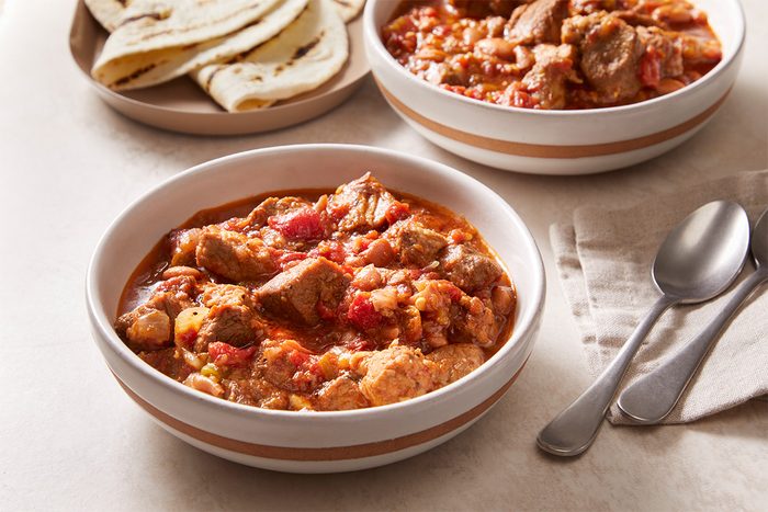 Two bowls of hearty stew with chunks of meat and tomatoes sit on a table next to a plate of folded flatbreads, two spoons, and a beige napkin.
