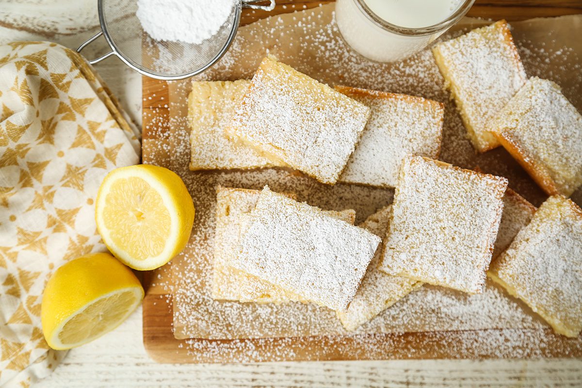 Rectangular lemon bars dusted with powdered sugar are arranged on a wooden board. A halved lemon, a metal sifter with powdered sugar, a glass of milk, and a patterned cloth are nearby.
