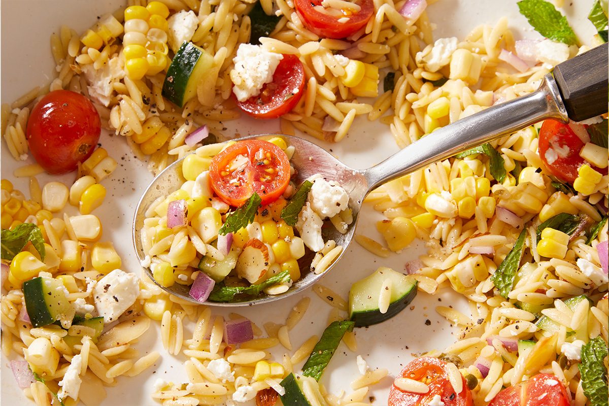 A close-up of a fresh orzo salad with cherry tomatoes, corn, cucumber, red onion, feta cheese, and herbs, served with a spoon.
