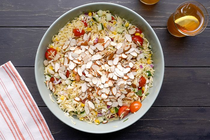 A bowl of orzo salad with cherry tomatoes, corn, herbs, and topped with sliced almonds sits on a dark wooden table next to a striped cloth and a glass of iced tea with a lemon slice.