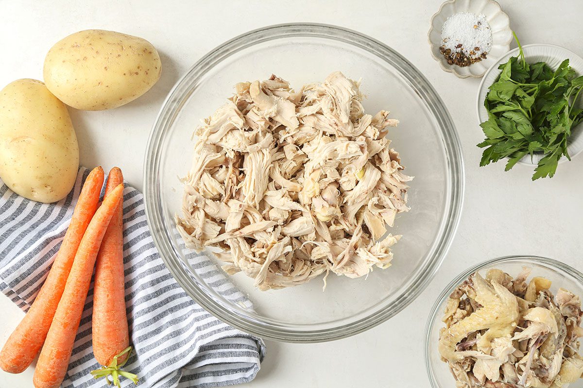 A glass bowl filled with shredded chicken sits on a white surface near whole potatoes, carrots, fresh greens, a striped towel, and a small bowl of salt and pepper.
