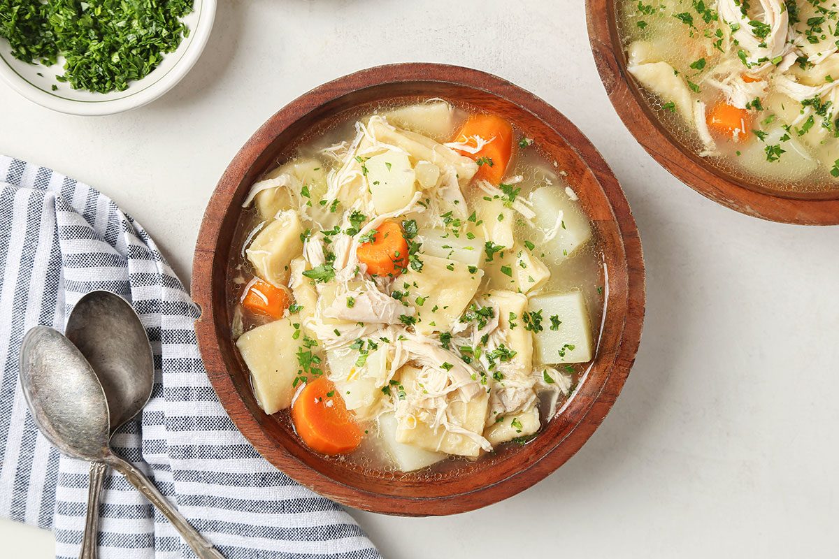 A wooden bowl of chicken noodle soup with shredded chicken, egg noodles, carrots, potatoes, and fresh herbs, beside a striped cloth, two spoons, and a small bowl of chopped parsley.