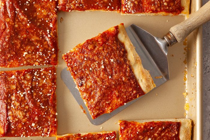 A metal spatula lifts a square slice of pizza with a thick, golden-brown crust and tomato sauce, surrounded by other pizza slices on a baking sheet.