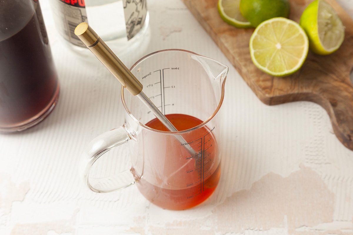 A glass measuring cup with reddish liquid and a stirring spoon inside. In the background, there are halved limes on a cutting board and bottles of liquid.