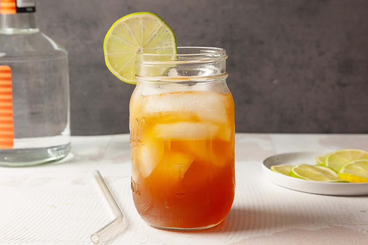 A mason jar filled with iced tea and ice cubes, garnished with a lime wheel, sits on a white surface with a clear straw nearby. In the background, there is a plate with lime slices and a bottle.