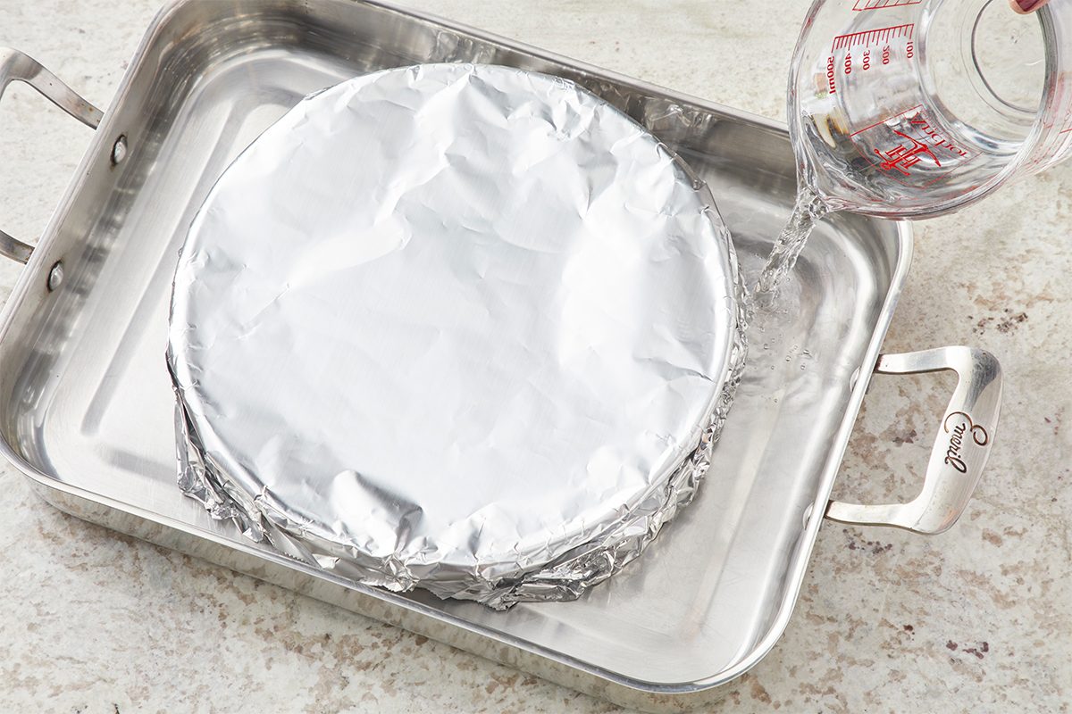 A foil-covered round pan sits inside a larger roasting pan as water is being poured from a glass measuring cup into the outer pan, preparing a water bath for baking.