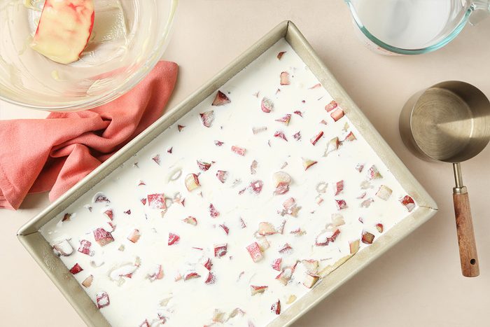 A baking tray filled with white liquid and sprinkled with small pieces of rhubarb sits on a countertop. Nearby are a glass mixing bowl with a pink cloth, a metal measuring cup, and a glass measuring jug.