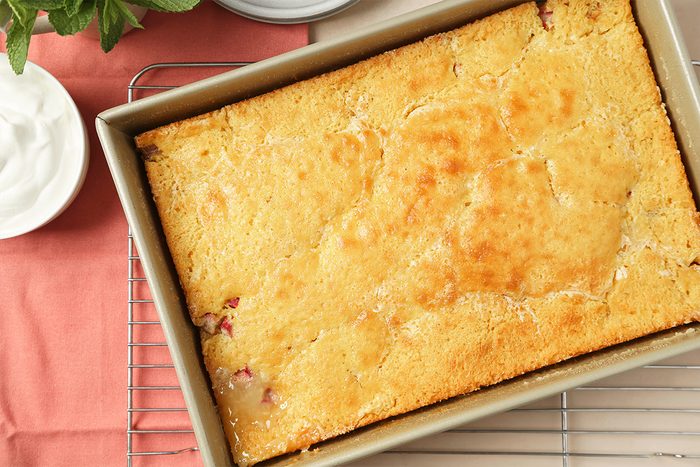 A golden-brown baked cake in a rectangular metal pan rests on a cooling rack, with a bowl of whipped cream and a pink napkin nearby.