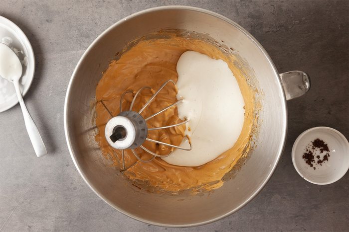 A metal mixing bowl contains a light brown batter, partially covered with a mound of white sugar. A whisk attachment is inside the bowl. Nearby are a spoon on a plate and a small bowl with some powder.