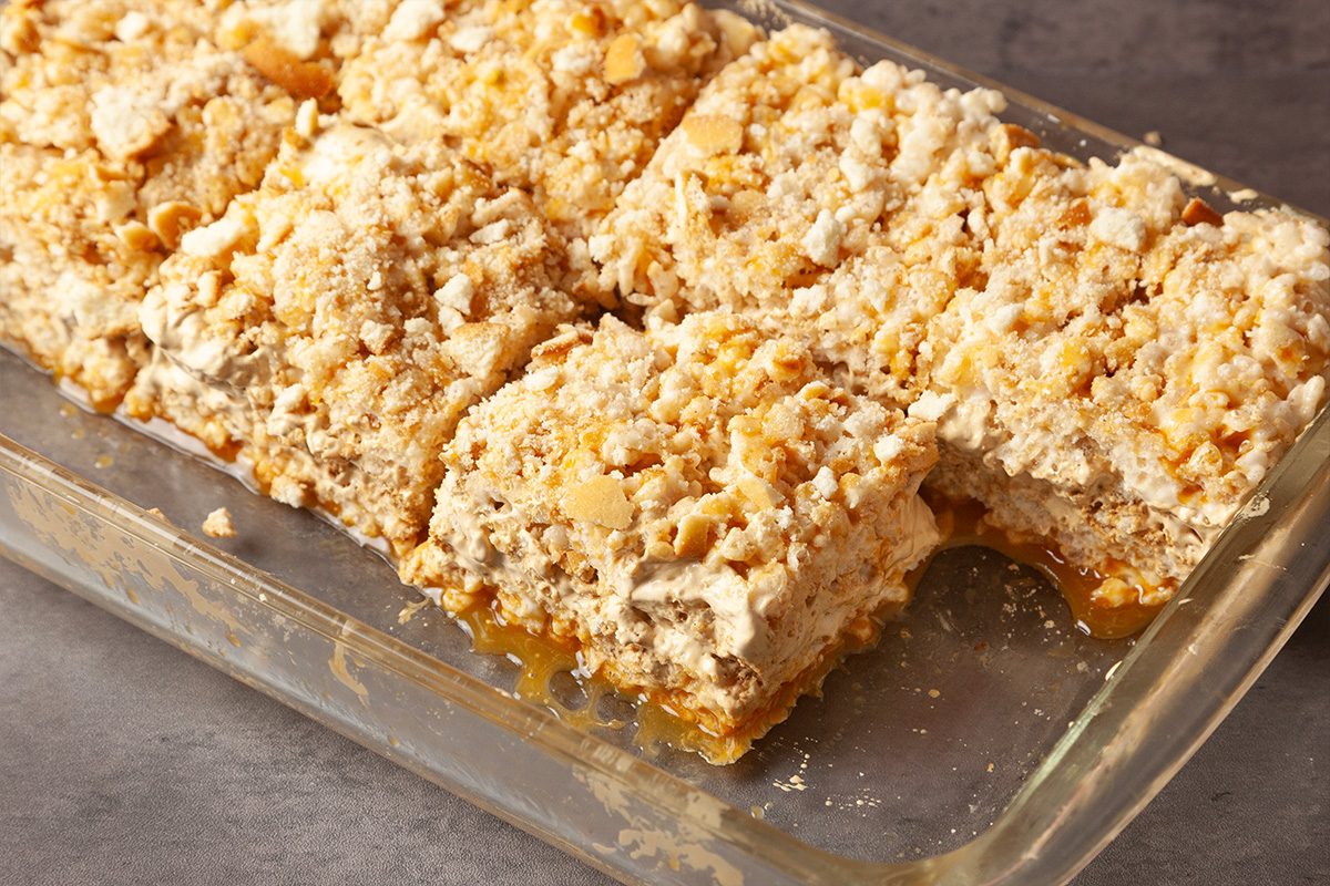A glass baking dish filled with six golden, crumbly dessert bars cut into squares, featuring a layered texture and a topping of crushed crumbs or nuts. The bars rest on a gray surface.