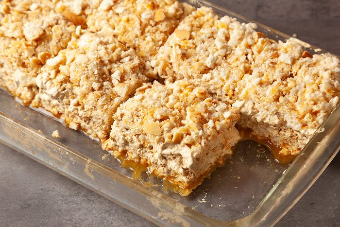 A glass baking dish filled with six golden, crumbly dessert bars cut into squares, featuring a layered texture and a topping of crushed crumbs or nuts. The bars rest on a gray surface.