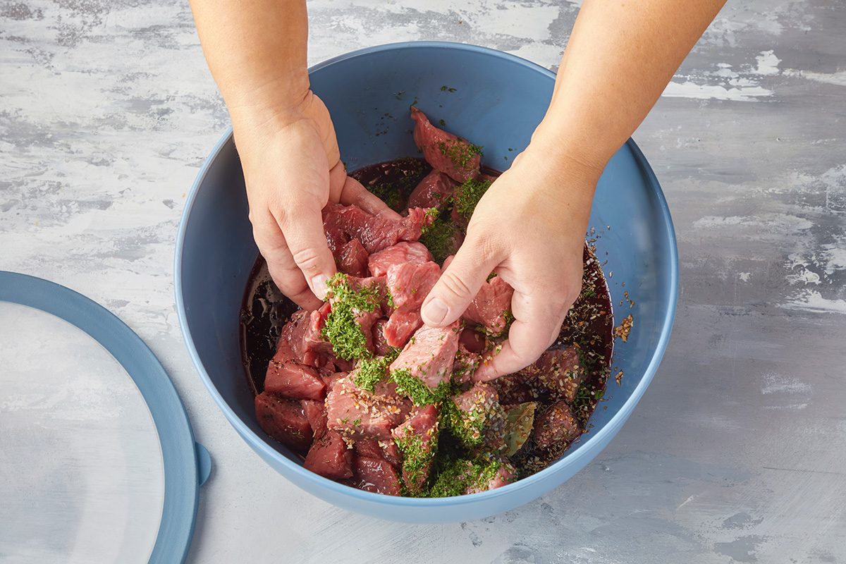 Hands mixing raw cubed meat with herbs and seasonings in a large blue bowl, preparing the meat for marinating. The bowl is placed on a light, textured surface.