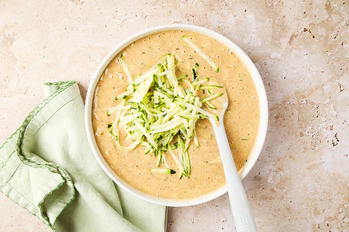 A bowl of creamy soup garnished with shredded zucchini, with a white spoon inside the bowl. A light green cloth napkin lies beside the bowl on a beige surface.