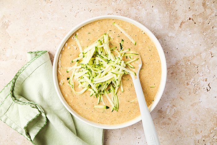 A bowl of creamy soup garnished with shredded zucchini, with a white spoon inside the bowl. A light green cloth napkin lies beside the bowl on a beige surface.