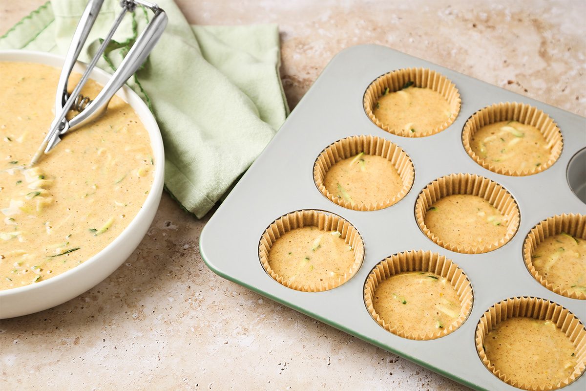 A bowl of muffin batter with two scoops beside a muffin tin lined with paper wrappers, some filled with batter, on a light countertop with a green cloth.