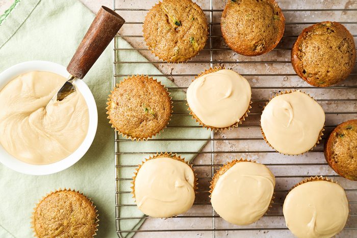 Cupcakes cooling on a wire rack, some plain and some topped with light brown frosting. A bowl of frosting with a spatula rests on the left, beside a green cloth.