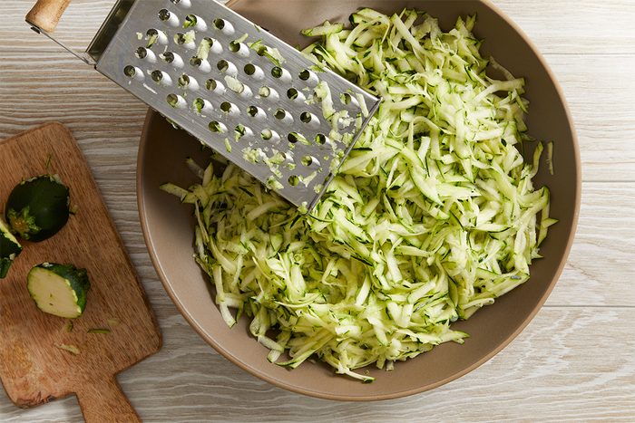 A bowl filled with freshly grated zucchini beside a metal grater, with zucchini ends placed on a wooden cutting board nearby, all set on a light wood surface.