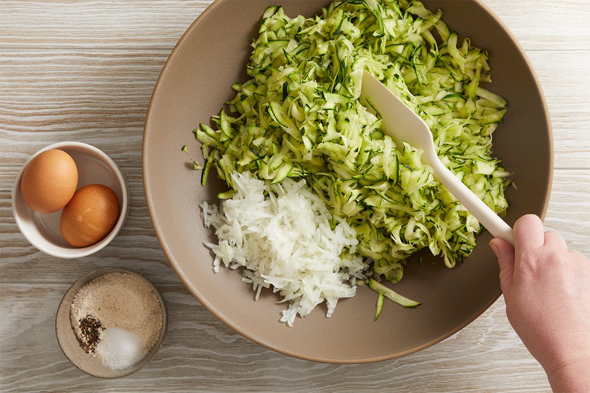 A hand mixes shredded zucchini and onion in a large bowl with a spatula. Nearby are two eggs in a small bowl and a bowl containing salt and pepper on a light wood surface.