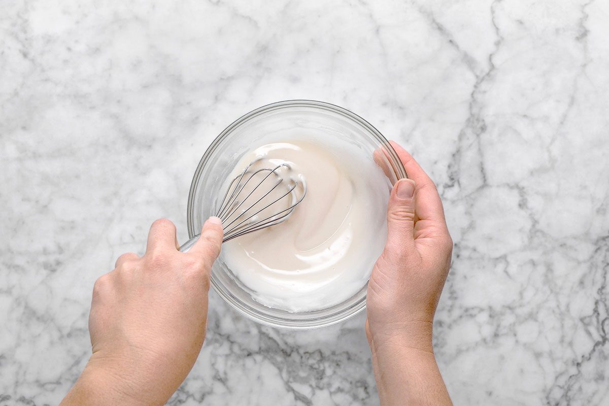 overhead shot of a person whisking a white, creamy mixture in a glass bowl on a marble countertop