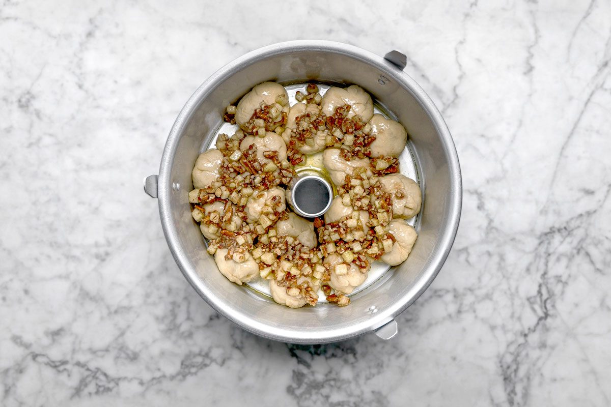 overhead shot of a round metal bundt pan filled with small dough balls topped with chopped nuts and brown sugar, sits on a white marble countertop