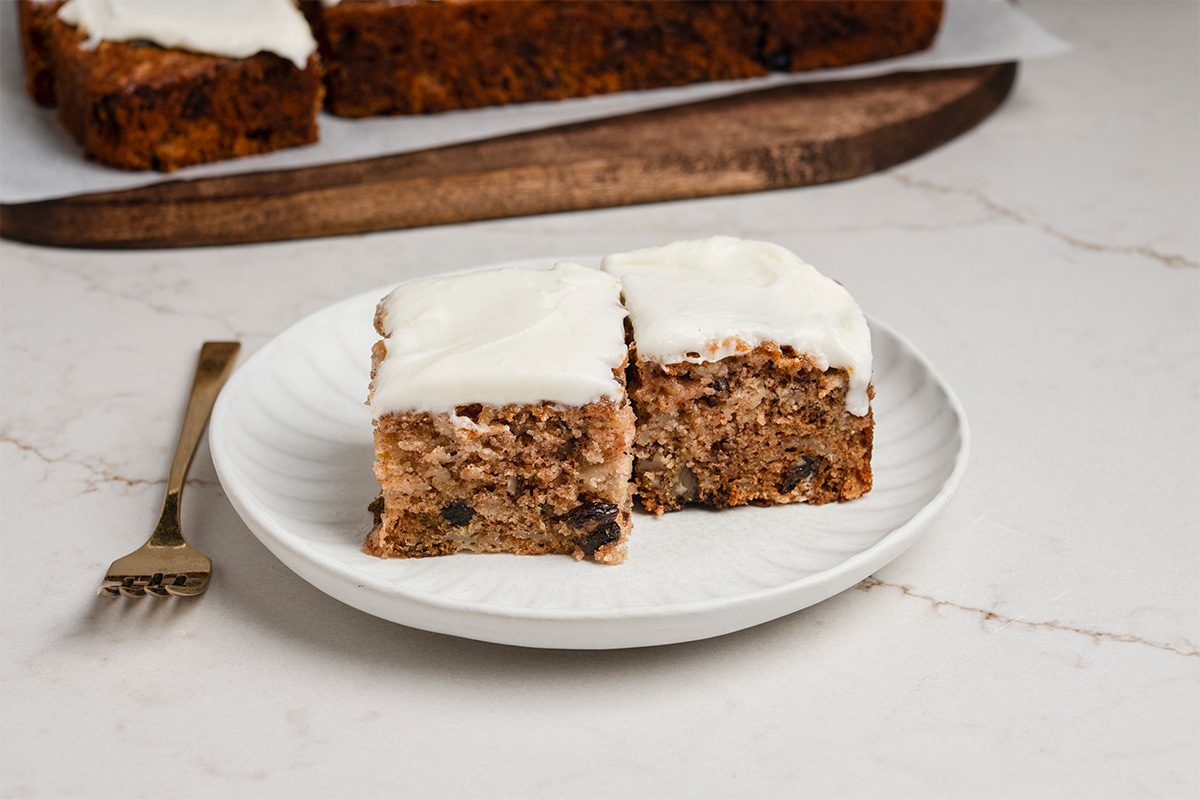 Two square pieces of spiced cake with white frosting on a white plate, next to a gold fork. More cake is visible in the background on a wooden board.
