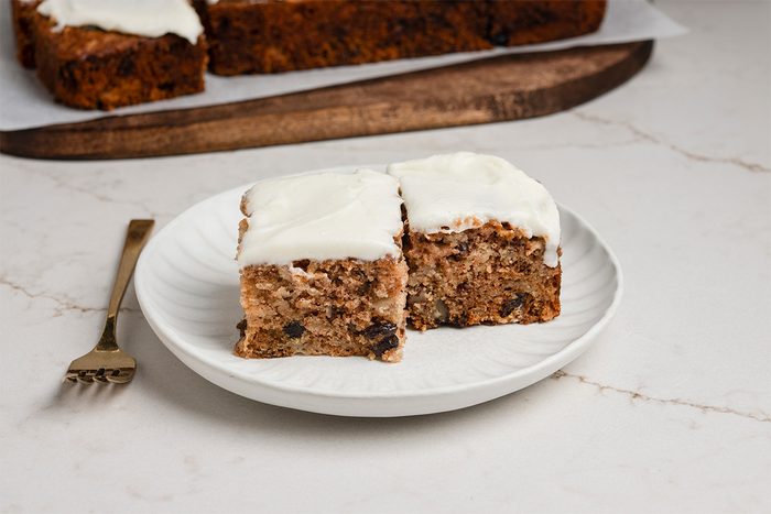 Two square pieces of spiced cake with white frosting on a white plate, next to a gold fork. More cake is visible in the background on a wooden board.