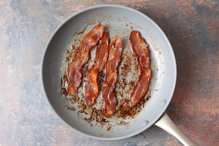 A frying pan containing three crispy cooked strips of bacon, with browned bits and grease visible on the pan’s surface.